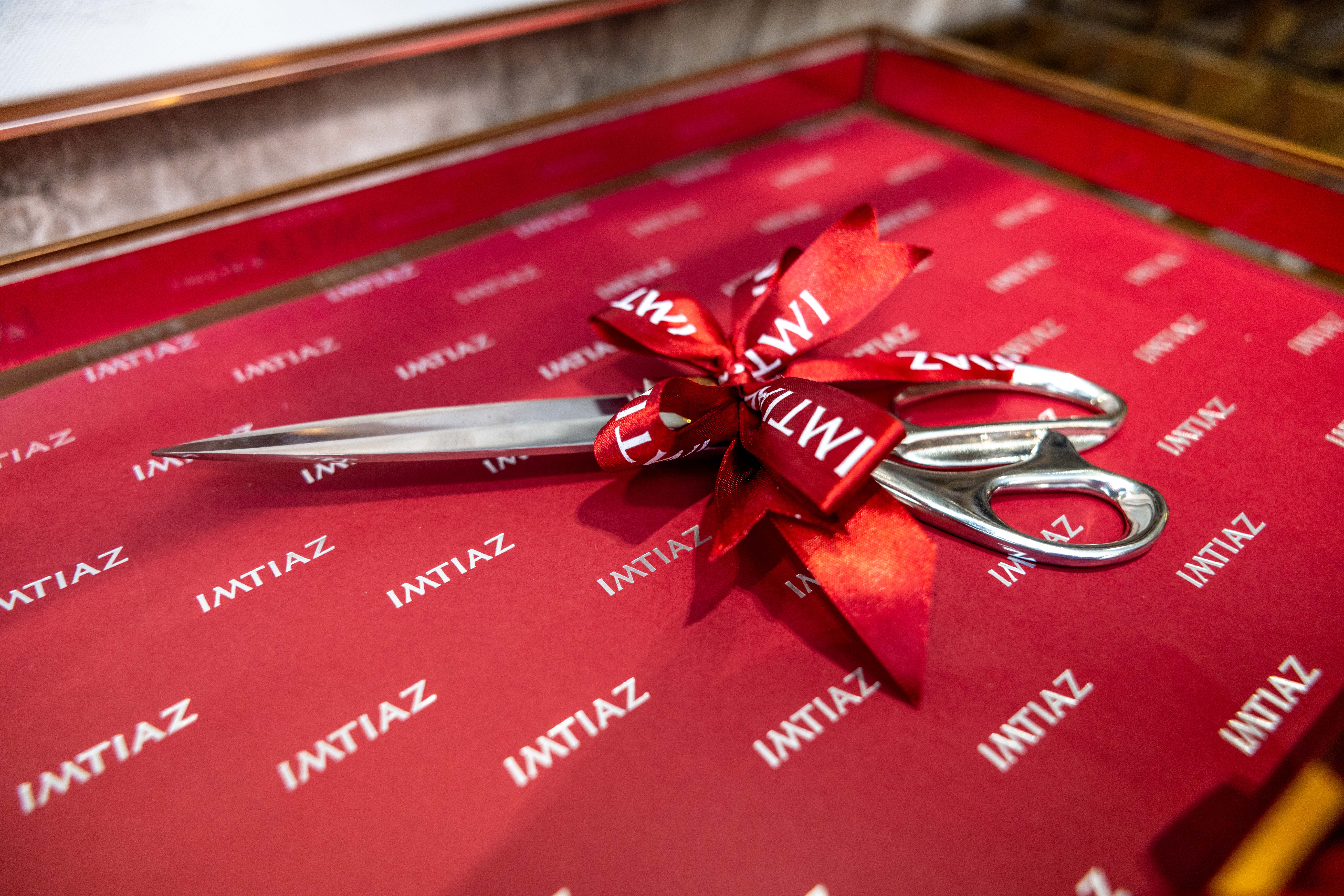 Events?Page=4 A pair of silver scissors with a red ribbon bow, placed on red paper featuring the word "IMTIAZ" repeatedly.