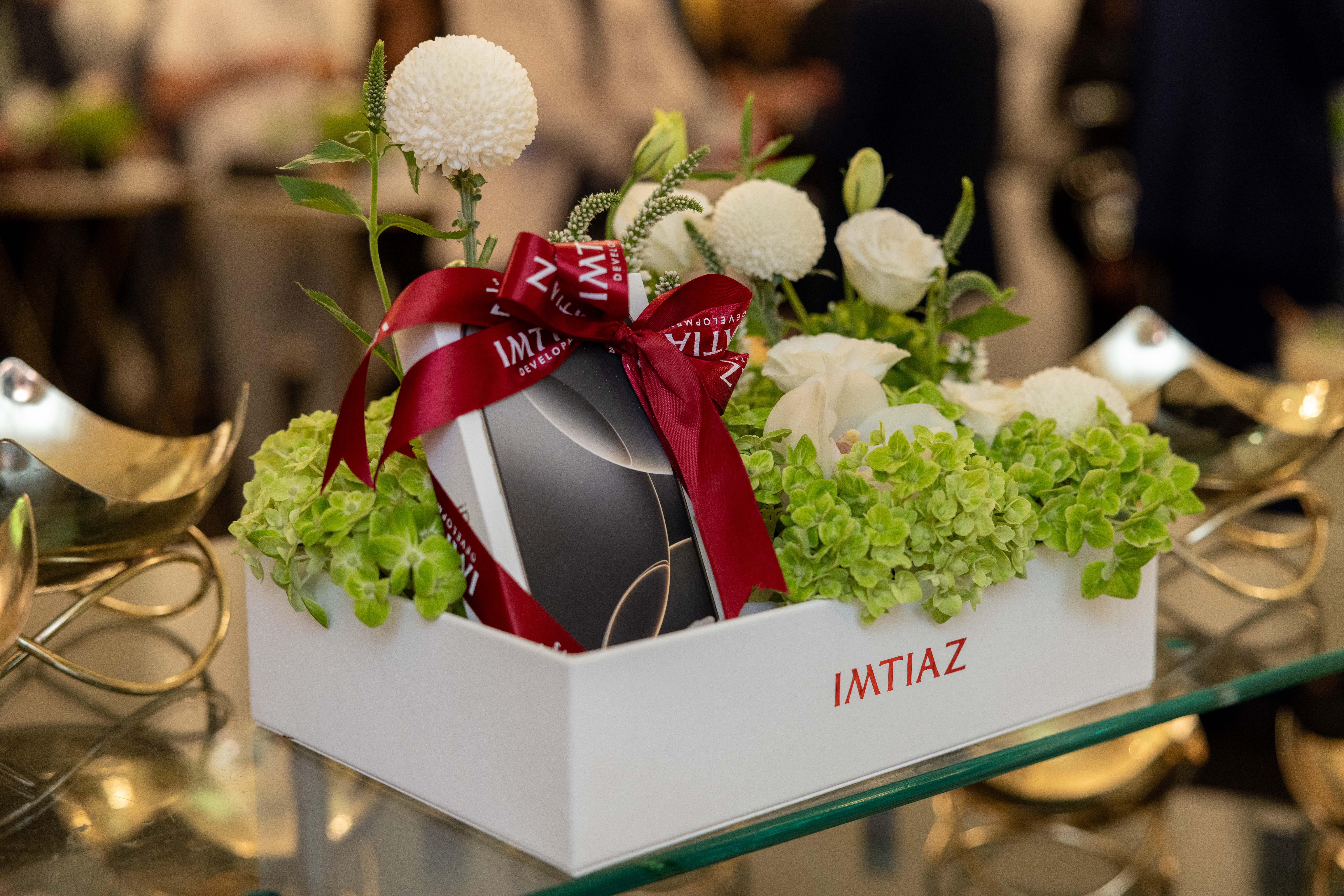 Events?Page=4 A decorative gift box with flowers and a ribbon, showcasing a small greenery-filled arrangement, labeled "IMTIAZ".