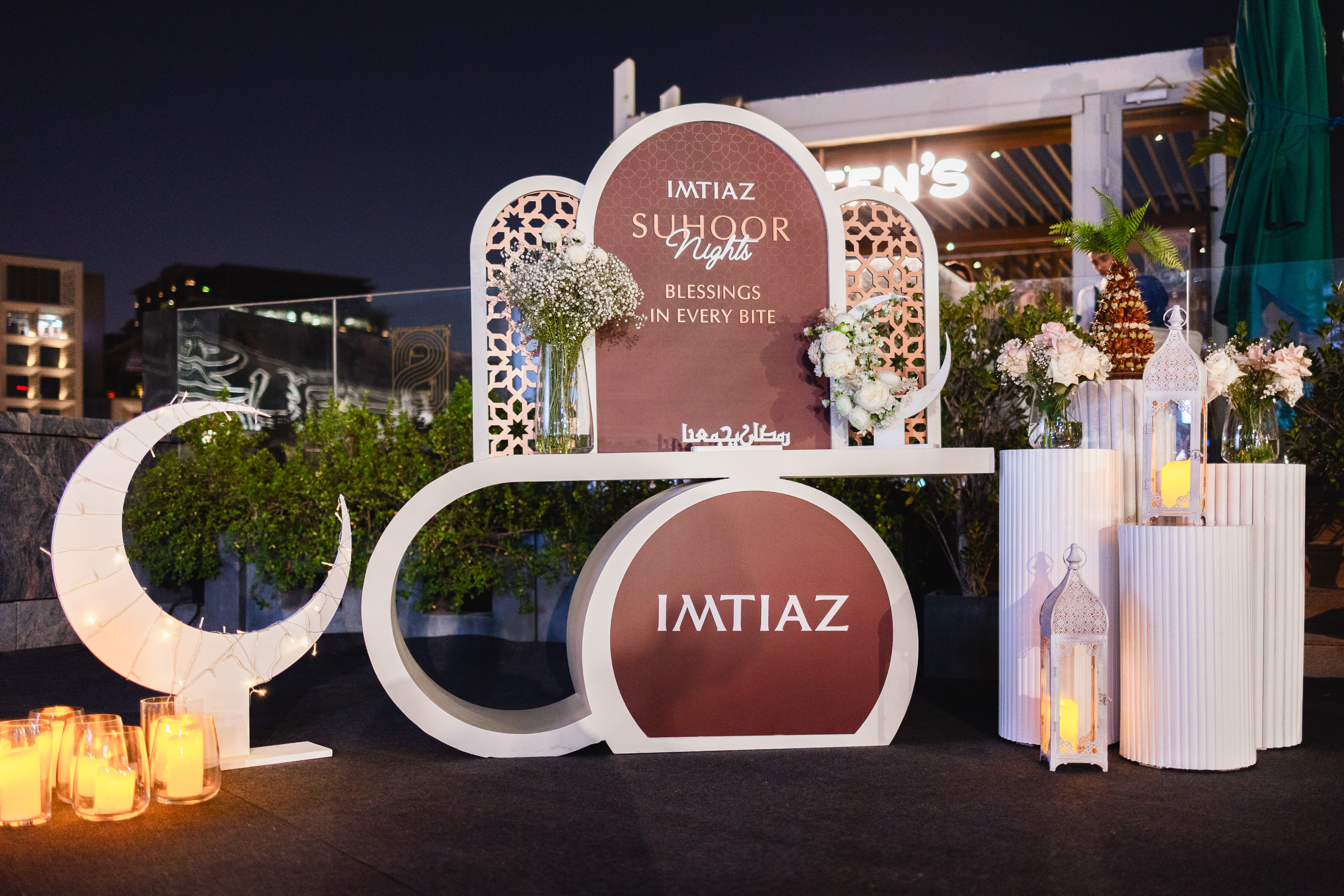 Imtiaz Ifthar Night A decorative setup with floral arrangements, lanterns, and a sign for "Imtiaz Suhoor Nights" in an evening setting.
