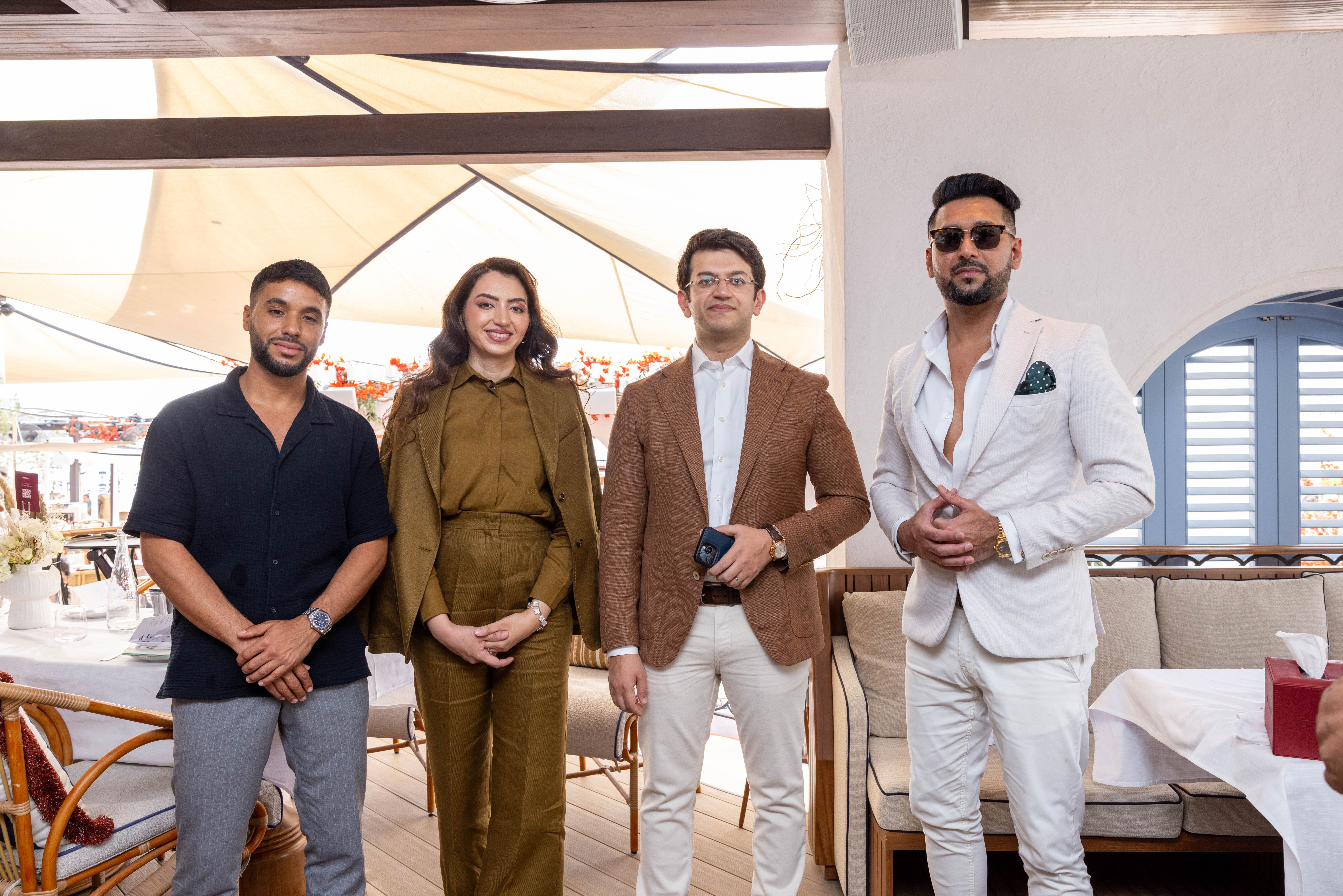 Events?Page=3 Four elegantly dressed individuals pose together indoors, showcasing a stylish atmosphere with dining chairs and decor.