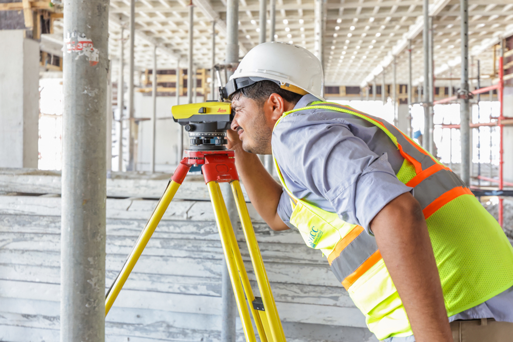 Pearl House By Imtiaz A construction worker in a hard hat uses a surveying instrument on a site with scaffolding and concrete structures.