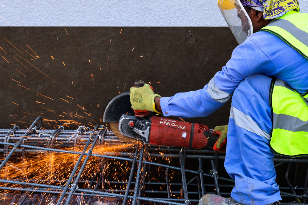 Pearl House By Imtiaz A construction worker in safety gear uses a cutting tool on steel reinforcement bars, creating sparks.