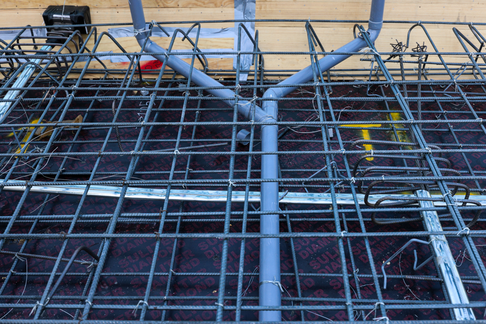 Pearl House By Imtiaz A top view of construction rebar grid with metal pipes and a wooden wall in the background, on a patterned surface.