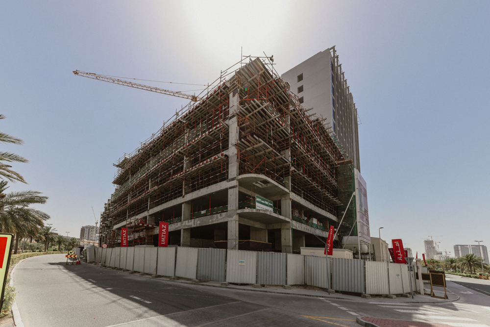 Pearl House By Imtiaz Construction site of a multi-story building with scaffolding, surrounded by barriers and palm trees under a clear sky.