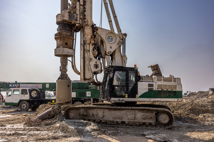 Beach Walk Residence A large drilling machine on a construction site, with soil and a green truck in the background under a clear sky.