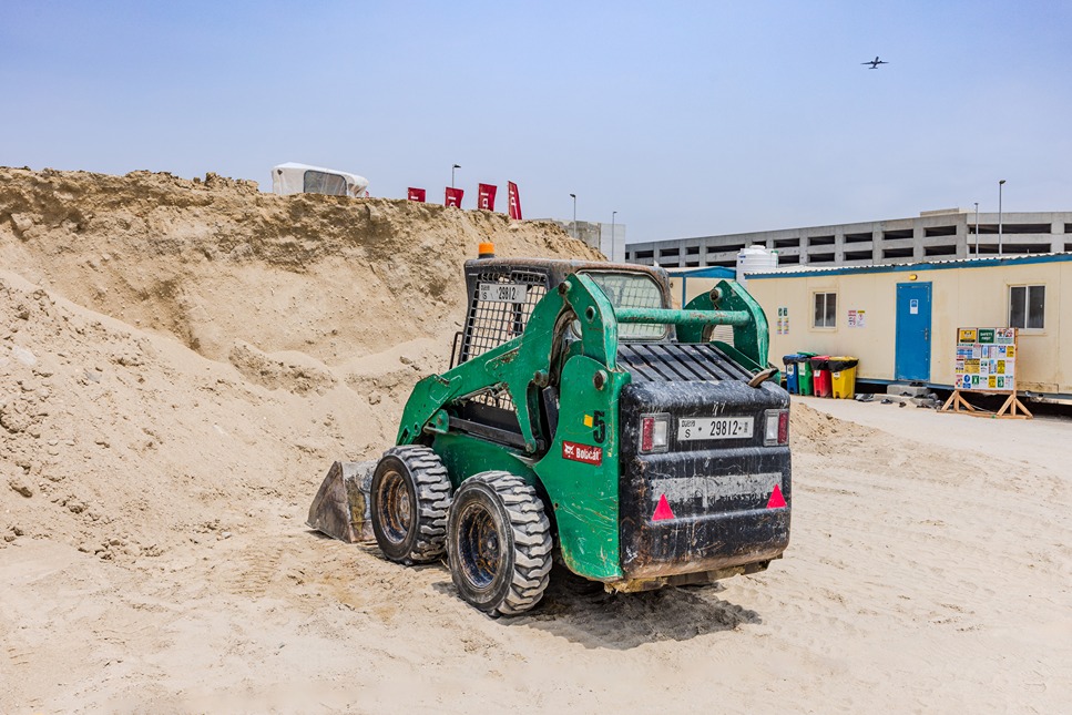 Beach Walk Residence A green skid steer loader parked on a sandy construction site with a building and trash bins in the background.