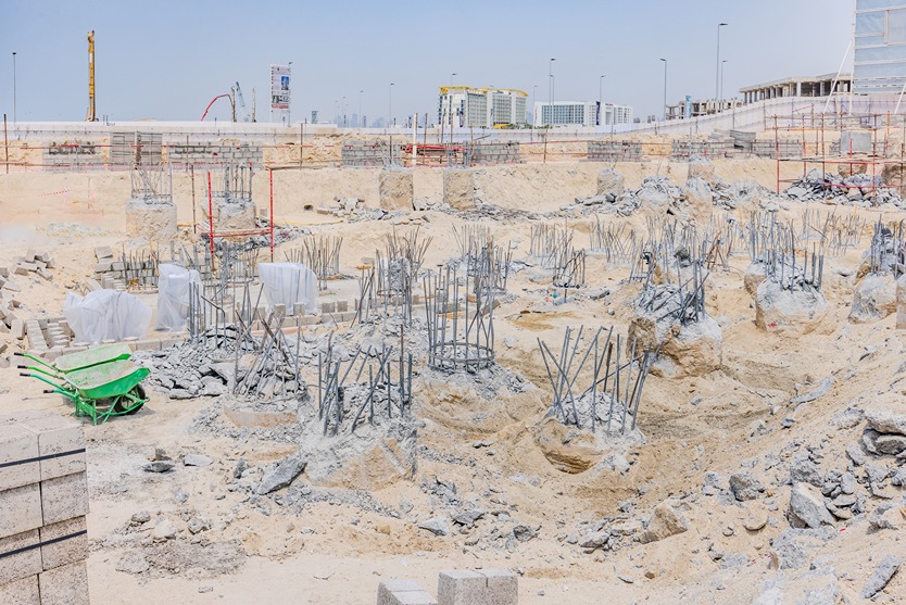 Beach Walk Residence Construction site with foundation pillars, rebar, and wheelbarrow amidst sandy terrain and building materials.