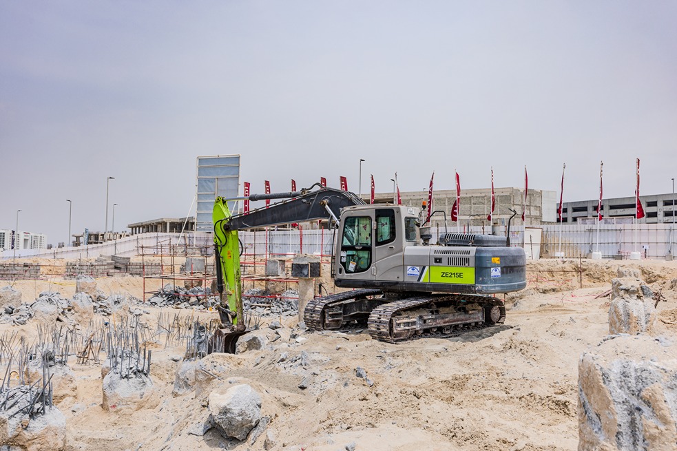 Beach Walk Residence A construction site featuring a large excavator among rocks and steel rebars, with a building under development in background.