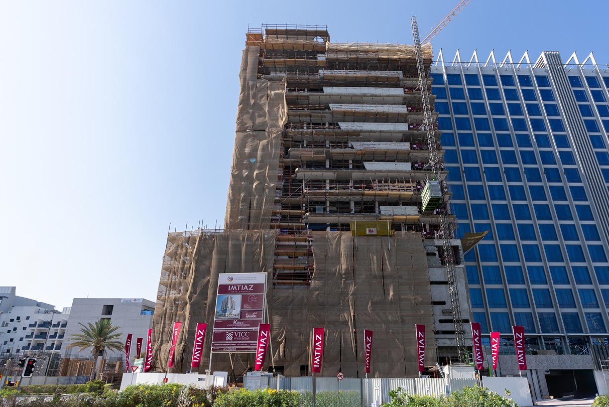 Pearl House By Imtiaz A tall building under construction, wrapped in netting, with a crane and vibrant banners in front.