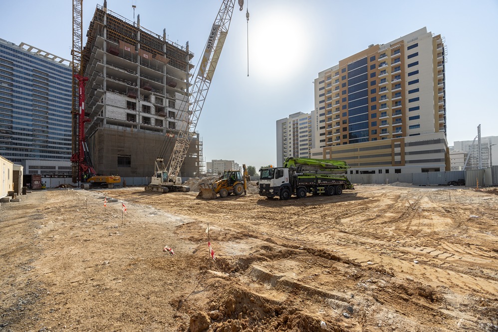 Cove Edition I By Imtiaz 1 A construction site featuring cranes, trucks, and ongoing building work under a bright sun backdrop.
