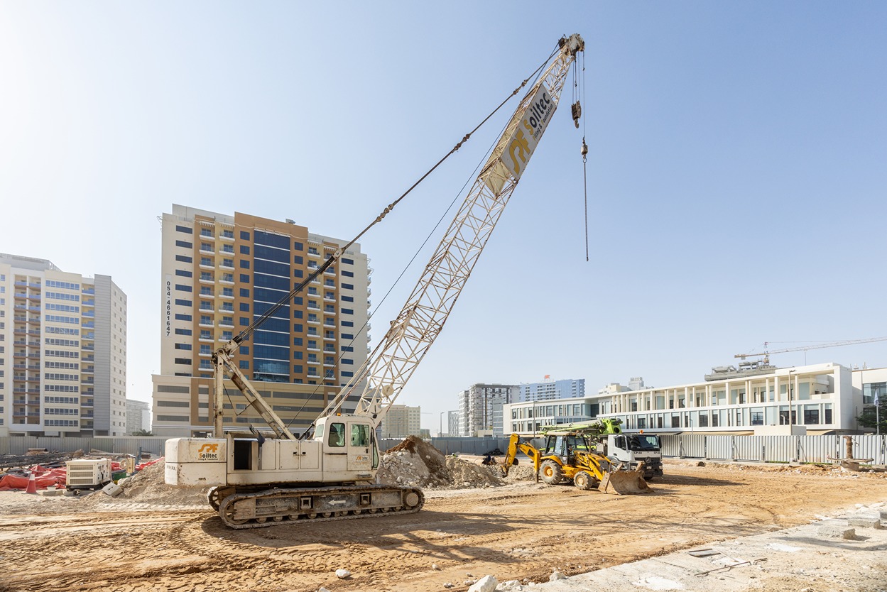 Cove Edition I By Imtiaz 1 A construction site featuring a crane, backhoe, and buildings in the background under a clear blue sky.