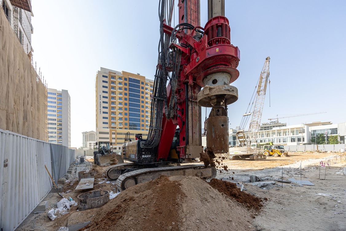 Cove Edition I By Imtiaz 1 Construction site with drilling machinery, buildings in the background, and dirt piles on the ground.