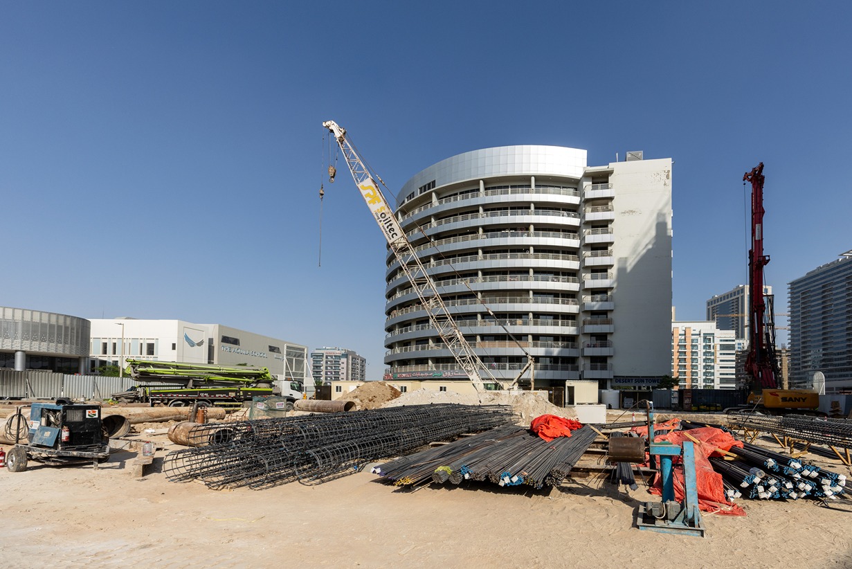 Cove Edition I By Imtiaz 1 Construction site with a crane, metal rebar piles, and a semi-finished building against a clear blue sky.