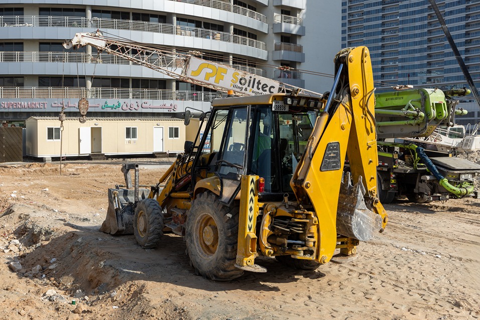 Cove Edition I By Imtiaz 1 A yellow backhoe loader parked on a construction site, with mobile offices and cranes in the background.