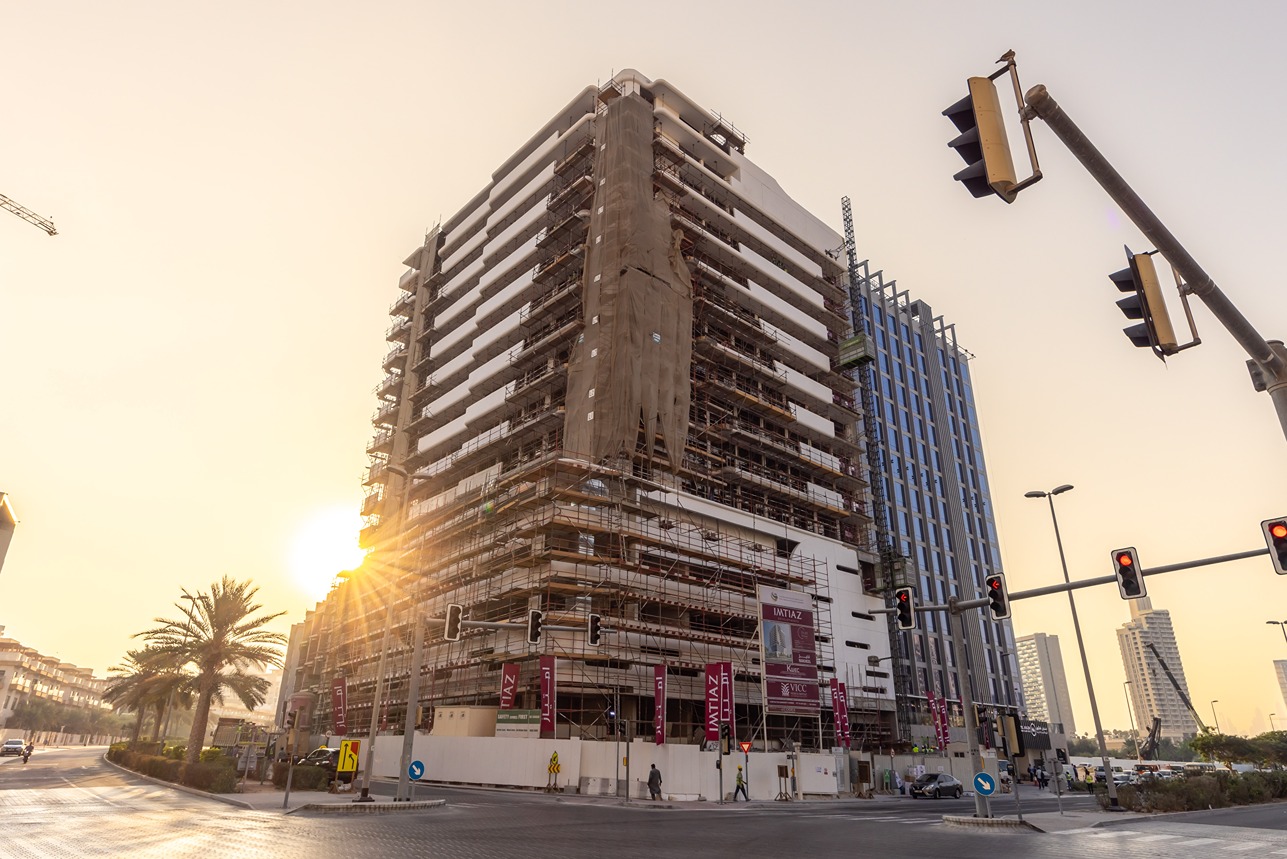 Pearl House By Imtiaz A partially constructed building with scaffolding and banners, street lights and palm trees, during sunset.