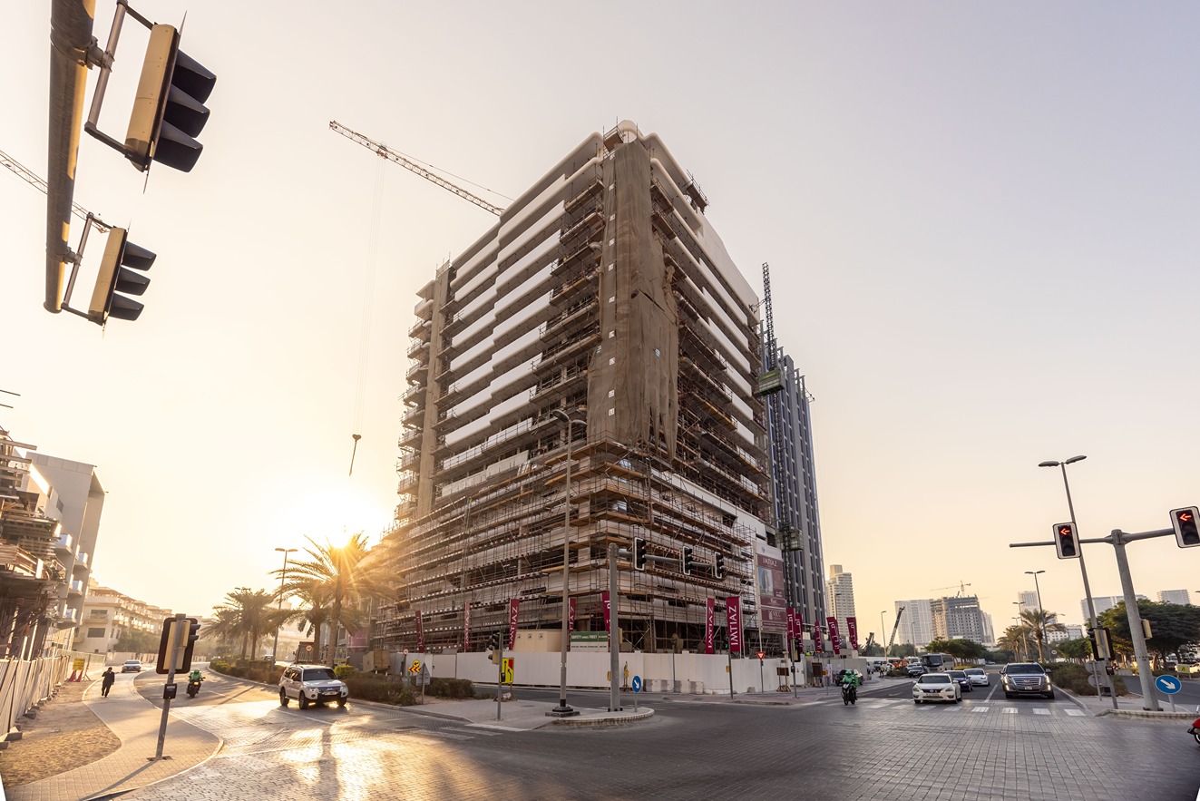 Pearl House By Imtiaz A large building under construction with scaffolding, traffic lights, and vehicles on a busy roadway at sunset.