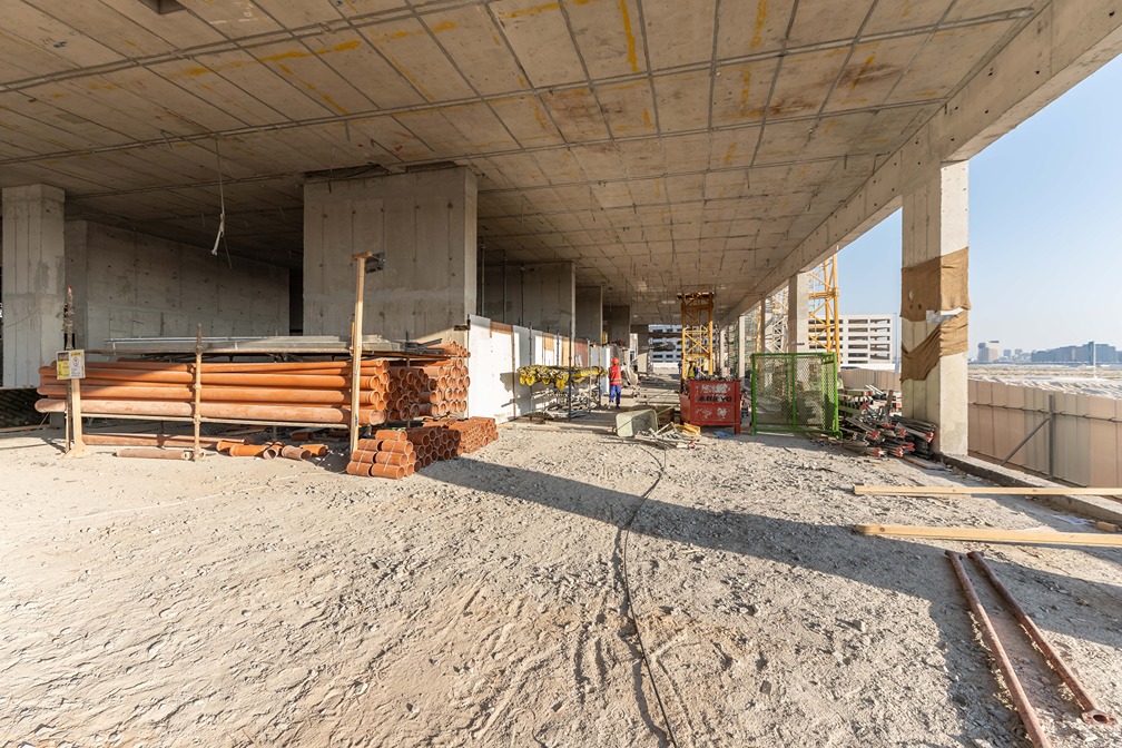 Beach Walk Residence Construction site interior with stacked materials, workers, and machinery under a concrete structure and bright sky.