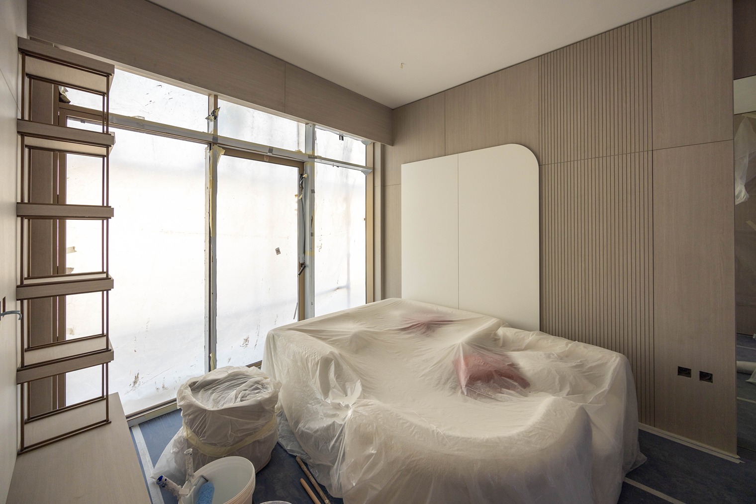 Pearl House By Imtiaz A partially renovated bedroom with a bed covered in plastic and a window, revealing construction work outside.