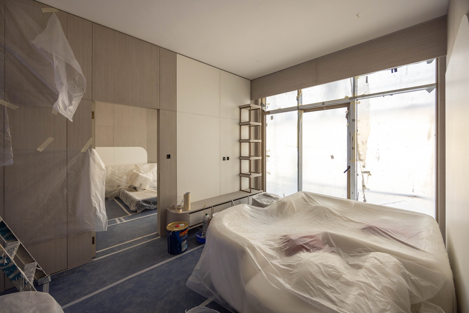 Pearl House By Imtiaz A partially furnished room with plastic coverings, a bed, and large windows, suggesting ongoing renovations.