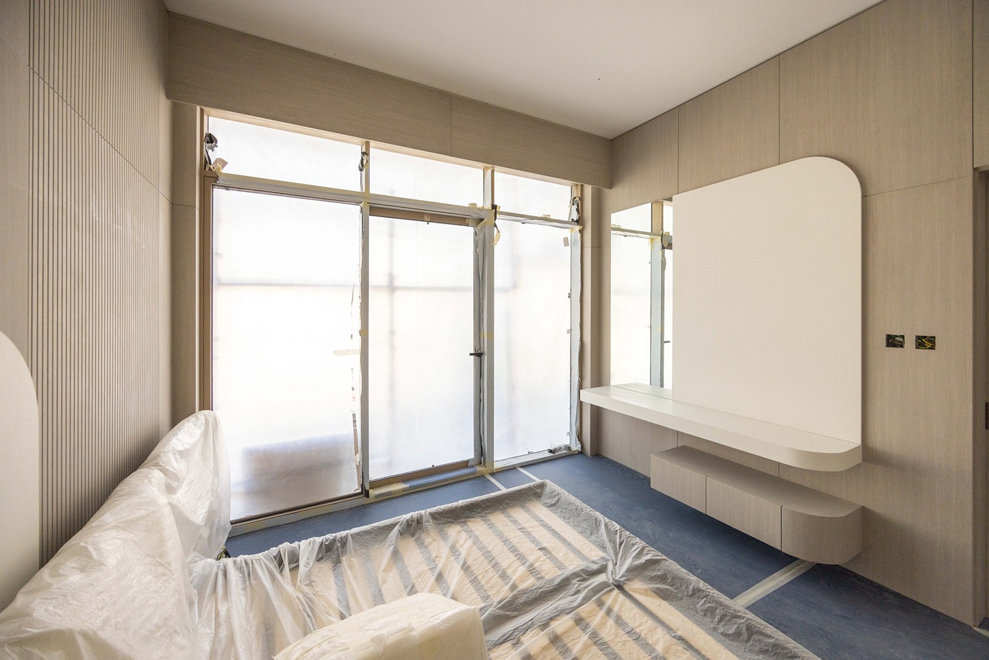 Pearl House By Imtiaz A partially decorated room with plastic covering furniture, large windows, and a modern wall-mounted cabinet.