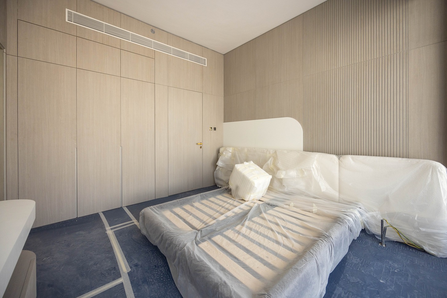 Pearl House By Imtiaz A minimally furnished room featuring a covered bed, wooden panels, and a bare blue floor under renovation.