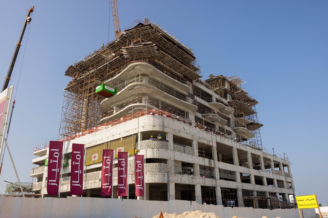 Beach Walk Residence A partially constructed building with scaffolding, construction workers, and banners in front. Clear blue sky above.