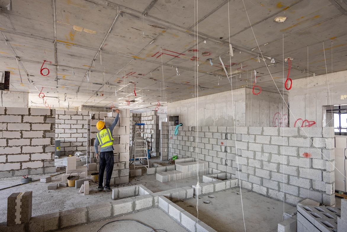 Beach Walk Residence A construction worker in a hard hat is building a wall with concrete blocks in an unfinished interior space.
