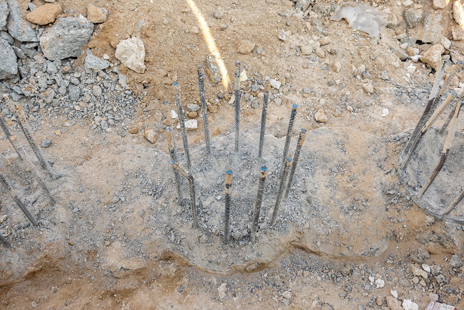 Cove Edition I By Imtiaz 1 Construction site with exposed concrete footings and protruding steel rebar, surrounded by dirt and gravel.