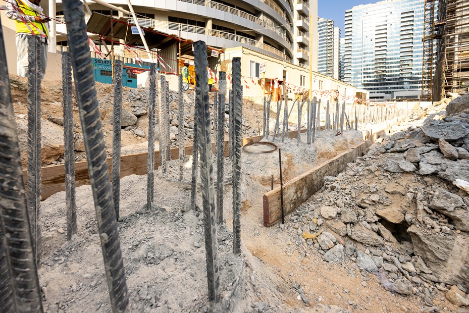 Cove Edition I By Imtiaz 1 Construction site showing rebar in concrete foundations with workers and buildings in the background.
