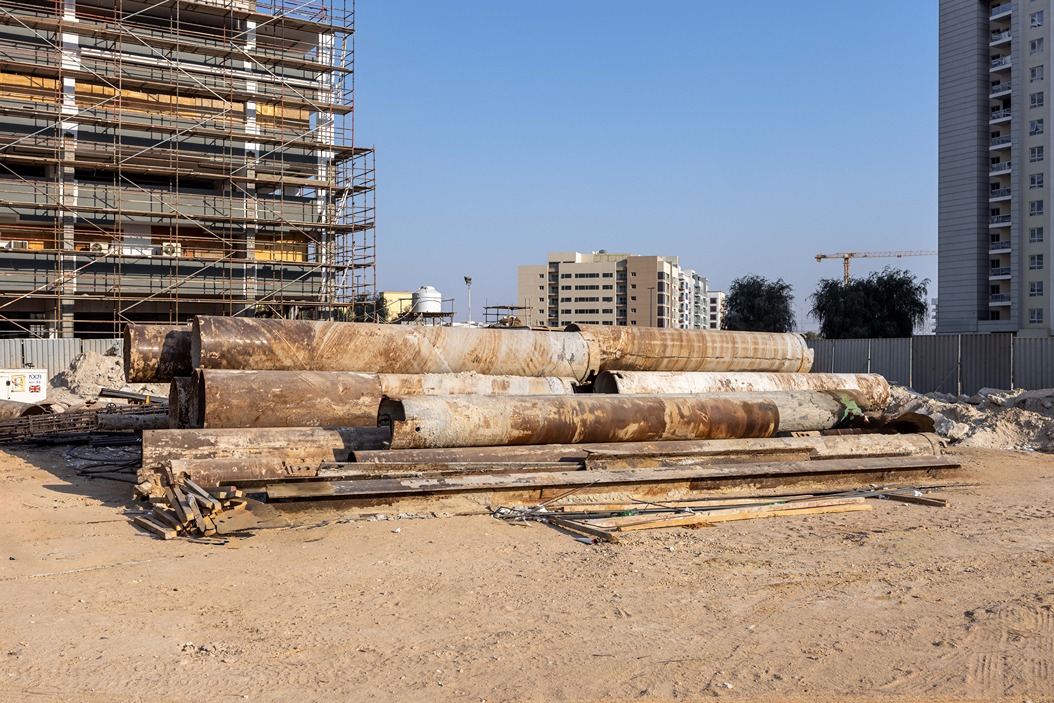 Cove Edition I By Imtiaz 1 Construction site displaying stacked metal pipes and scaffolding near buildings under development. Clear blue sky.