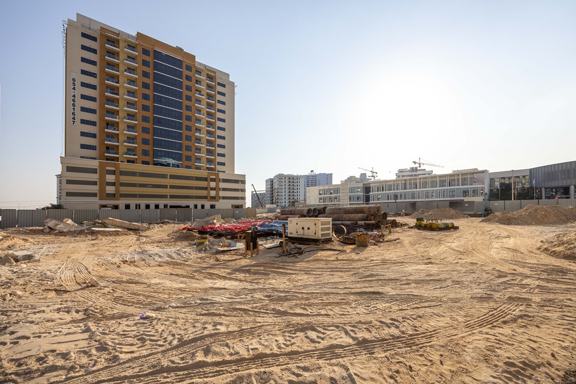Cove Edition I By Imtiaz 1 Construction site with a tall building, machinery, and scattered materials on a sandy landscape under a clear sky.