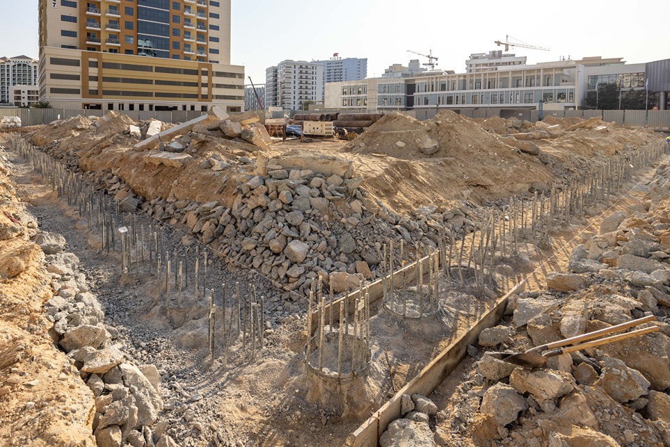 Cove Edition I By Imtiaz 1 Construction site with earth mounds, exposed rebar, and piles of rocks, surrounded by buildings and cranes in the background.