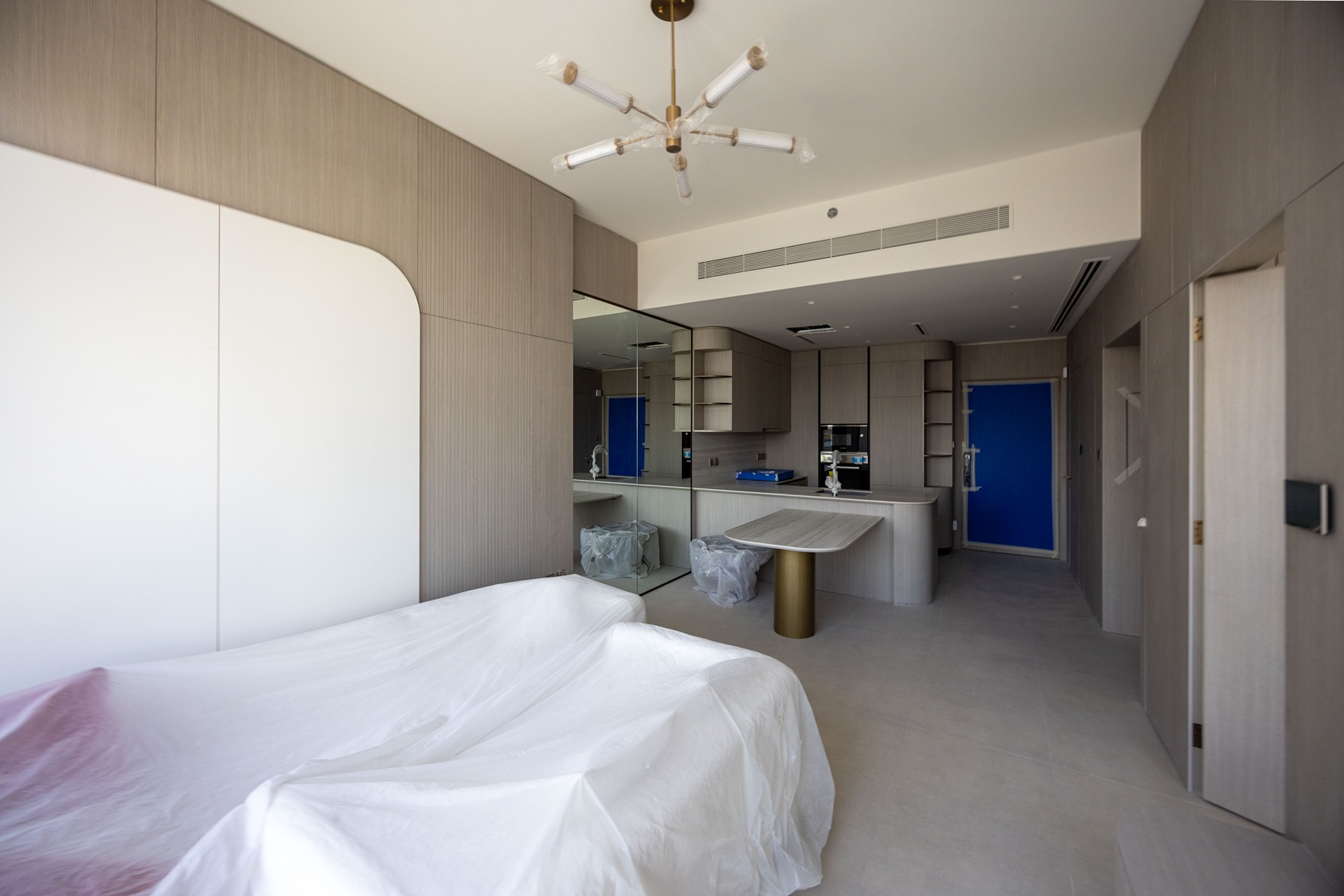 Pearl House By Imtiaz A modern, partially furnished room featuring a kitchen, dining table, and a covered sofa. Soft neutral tones dominate.