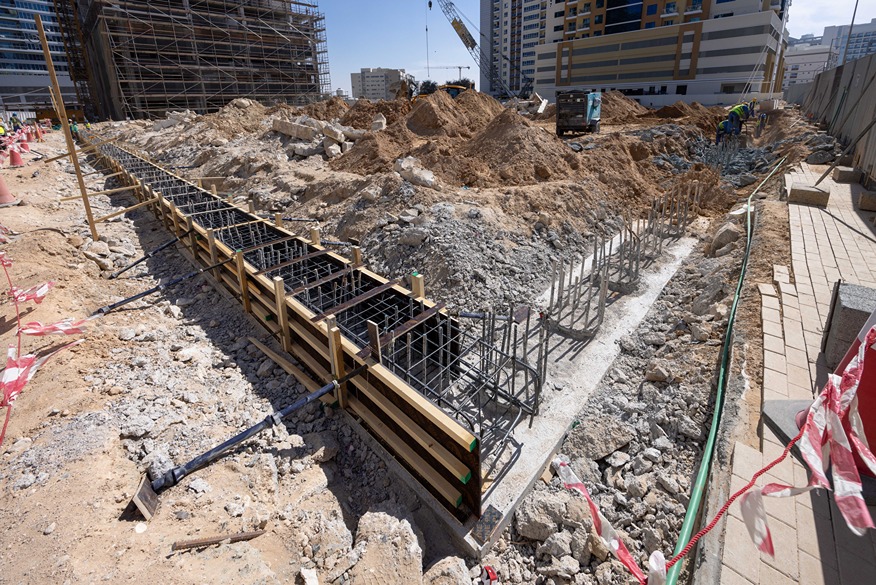 Cove Edition I By Imtiaz 1 Construction site with excavated ground, rebar structures, and wooden forms, surrounded by machinery and scaffolding.