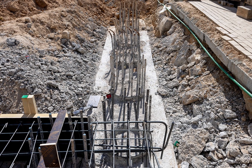 Cove Edition I By Imtiaz 1 A construction site showing rebar structures set in a trench surrounded by gravel and soil, with machinery nearby.