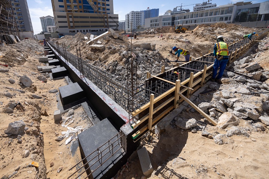Cove Edition I By Imtiaz 1 Construction site with workers assembling a concrete foundation amid rubble and sand, surrounded by buildings.