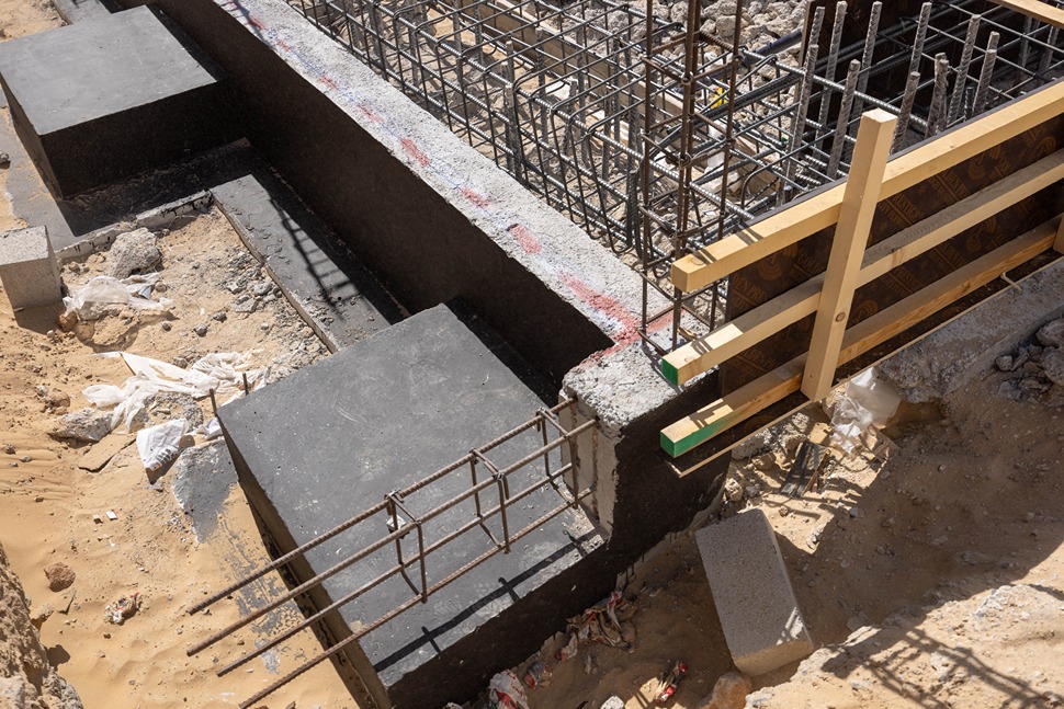 Cove Edition I By Imtiaz 1 Construction site featuring concrete foundations, rebar, wood forms, and scattered debris on sandy ground.