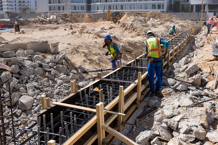 Cove Edition I By Imtiaz 1 Construction workers in helmets and vests work on a building site, assembling a concrete foundation framework.