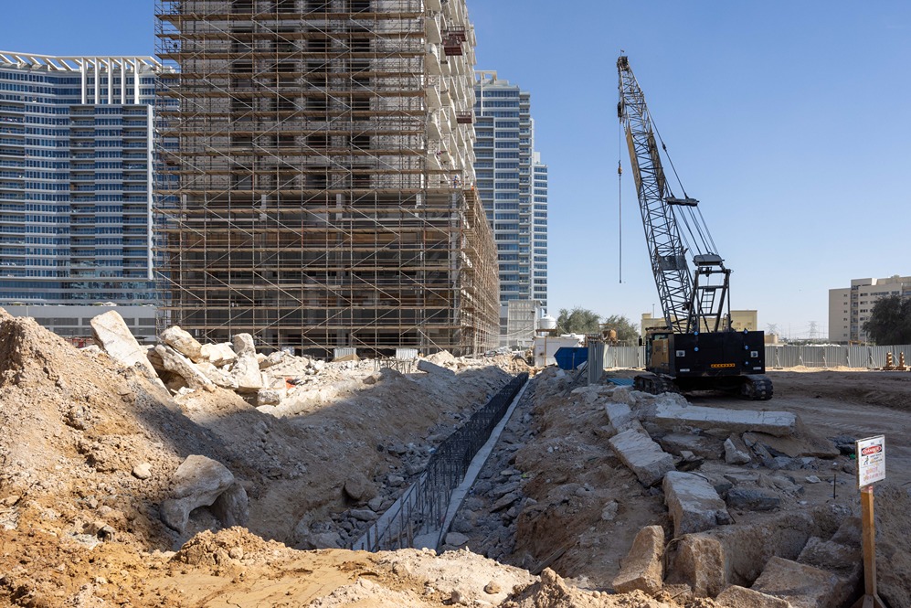 Cove Edition I By Imtiaz 1 Construction site with a tall building under scaffolding, a crane, and debris scattered around. Clear blue sky.