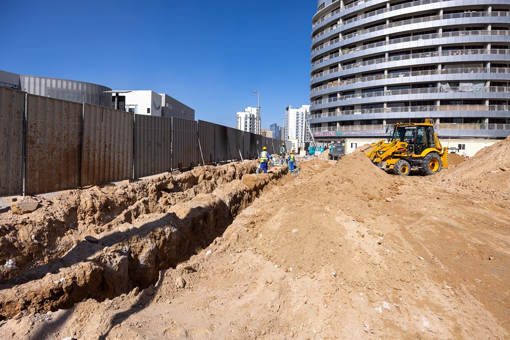 Cove Edition I By Imtiaz 1 Construction site with trench excavation, heavy machinery, and workers near a multi-story building. Clear blue sky.