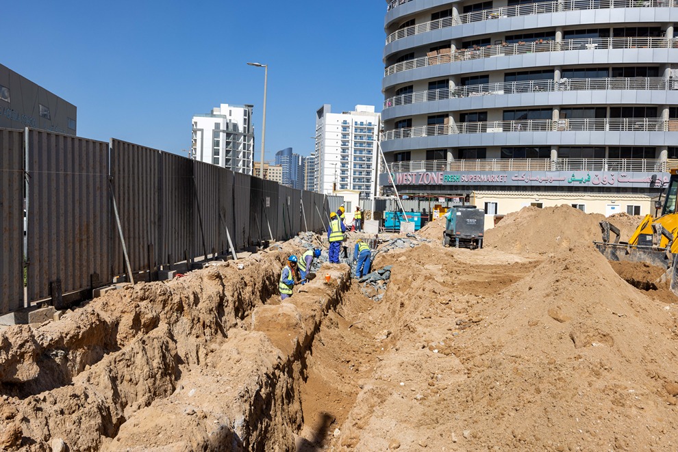 Cove Edition I By Imtiaz 1 Construction site with workers in safety gear digging trenches, surrounded by dirt mounds and modern buildings.