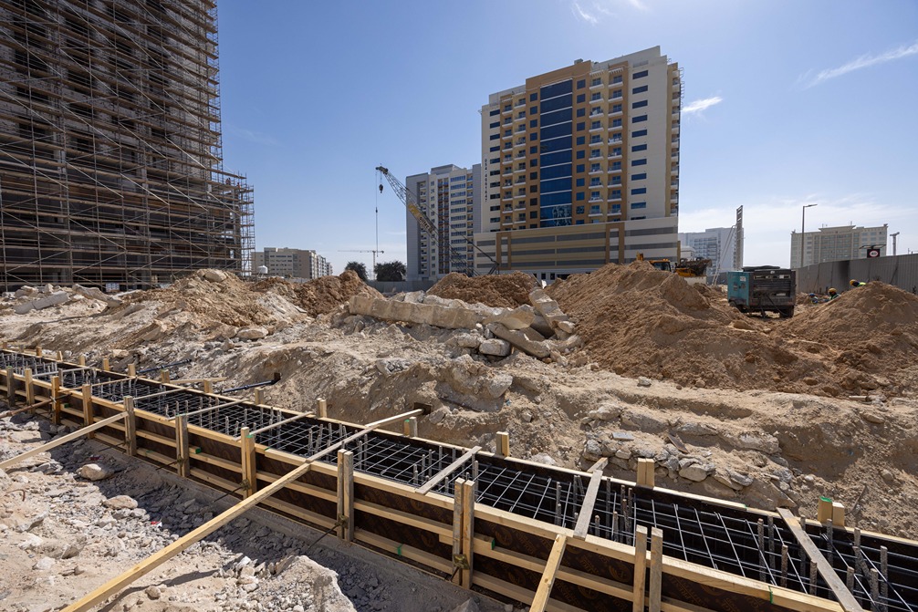 Cove Edition I By Imtiaz 1 Construction site with piles of dirt and concrete forms, high-rise buildings in the background under a clear sky.