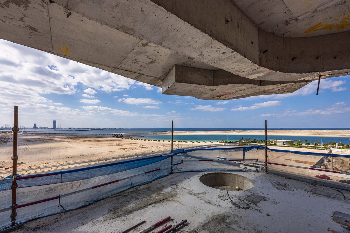 Beach Walk Residence View from a construction site overlooking a sandy beach, water, and distant buildings under a cloudy sky.