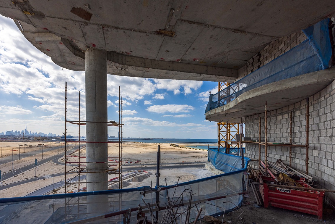 Beach Walk Residence View from a construction site showing scaffolding, a coastal landscape, and city skyline in the distance.