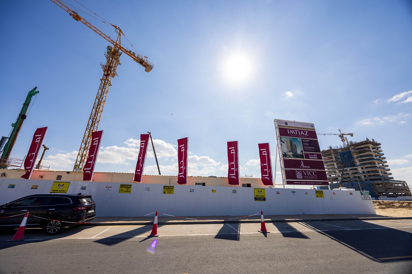Beach Walk Iii By Imtiaz Construction site with cranes, banners, safety signs, and a partially built building under a clear blue sky.