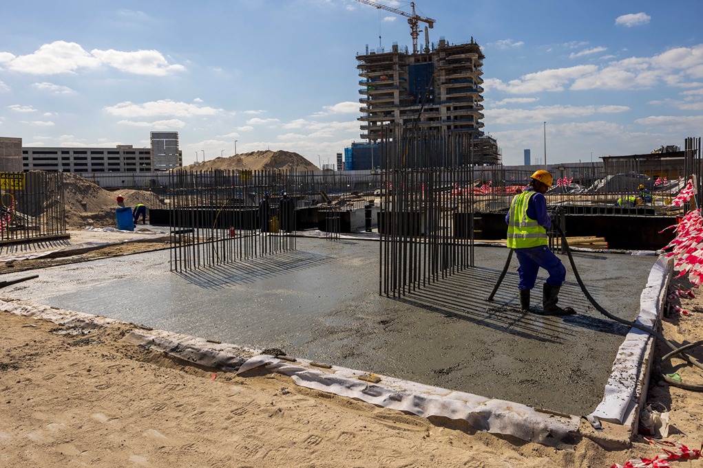 Beach Walk Iii By Imtiaz Construction site with a worker pouring concrete, steel rebar structures, and a developing building in the background.