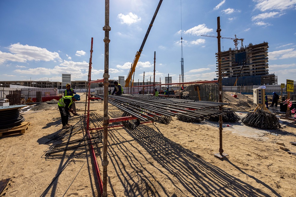 Beach Walk Iii By Imtiaz Construction site with workers, scaffolding, and steel rods under a clear sky and a building in progress.