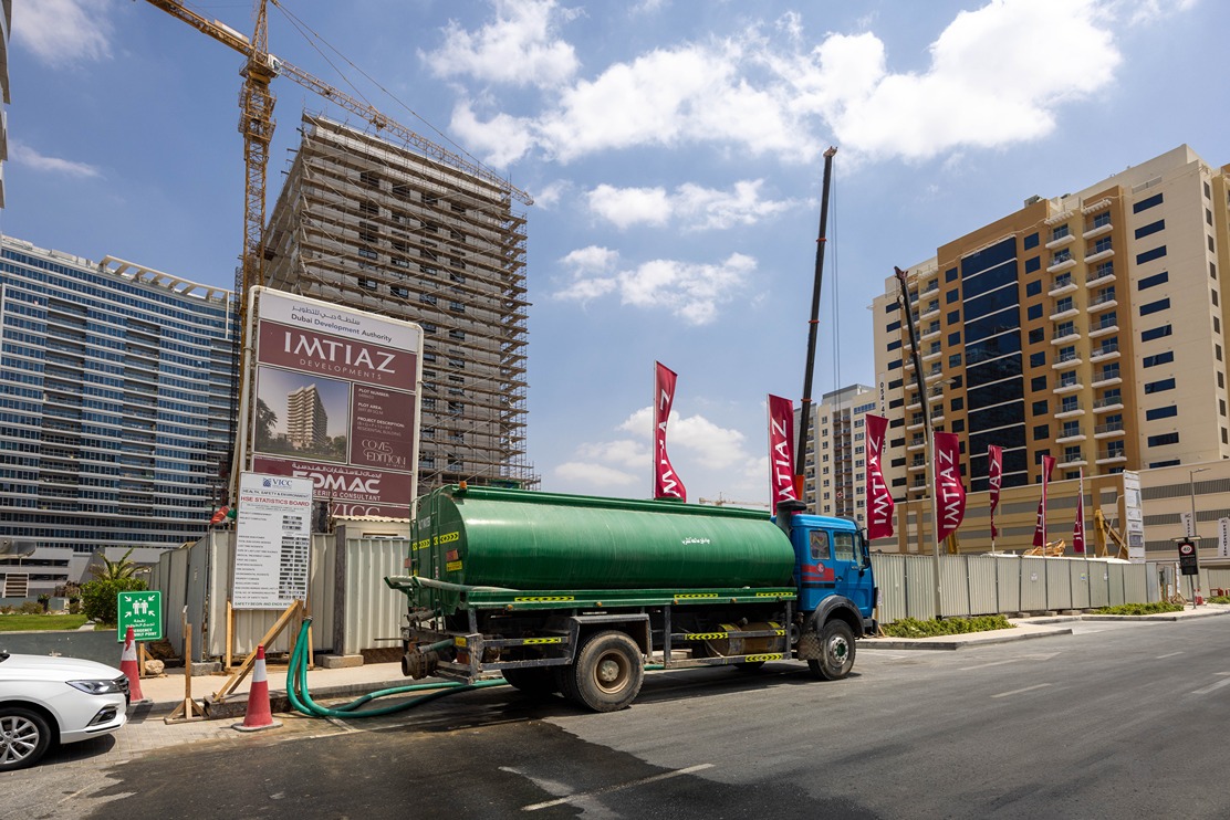 Cove Edition I By Imtiaz 1 A blue truck with a green water tank is parked near a construction site with tall buildings and banners.