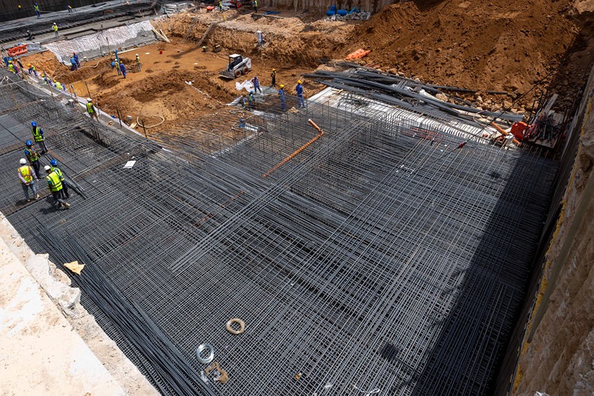 Cove Edition I By Imtiaz 1 Construction site showing workers on a rebar framework with earthmoving equipment and excavated soil in the background.