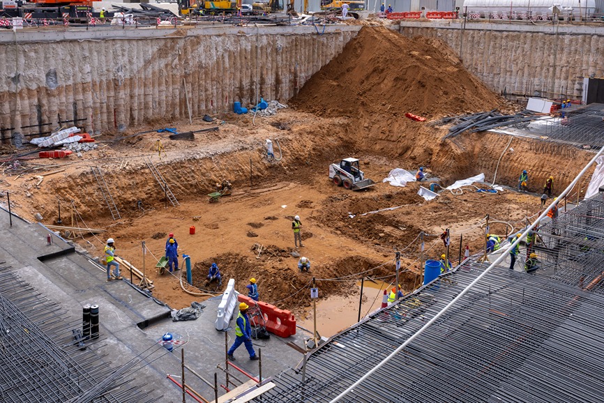 Cove Edition I By Imtiaz 1 Construction site showing workers, heavy machinery, and a large excavation surrounded by safety barriers and materials.